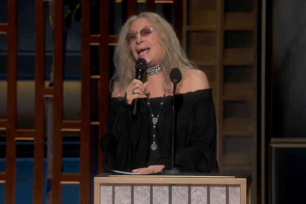 Barbra Streisand speaking at a microphone while wearing glasses and a black, off-the-shoulder top.